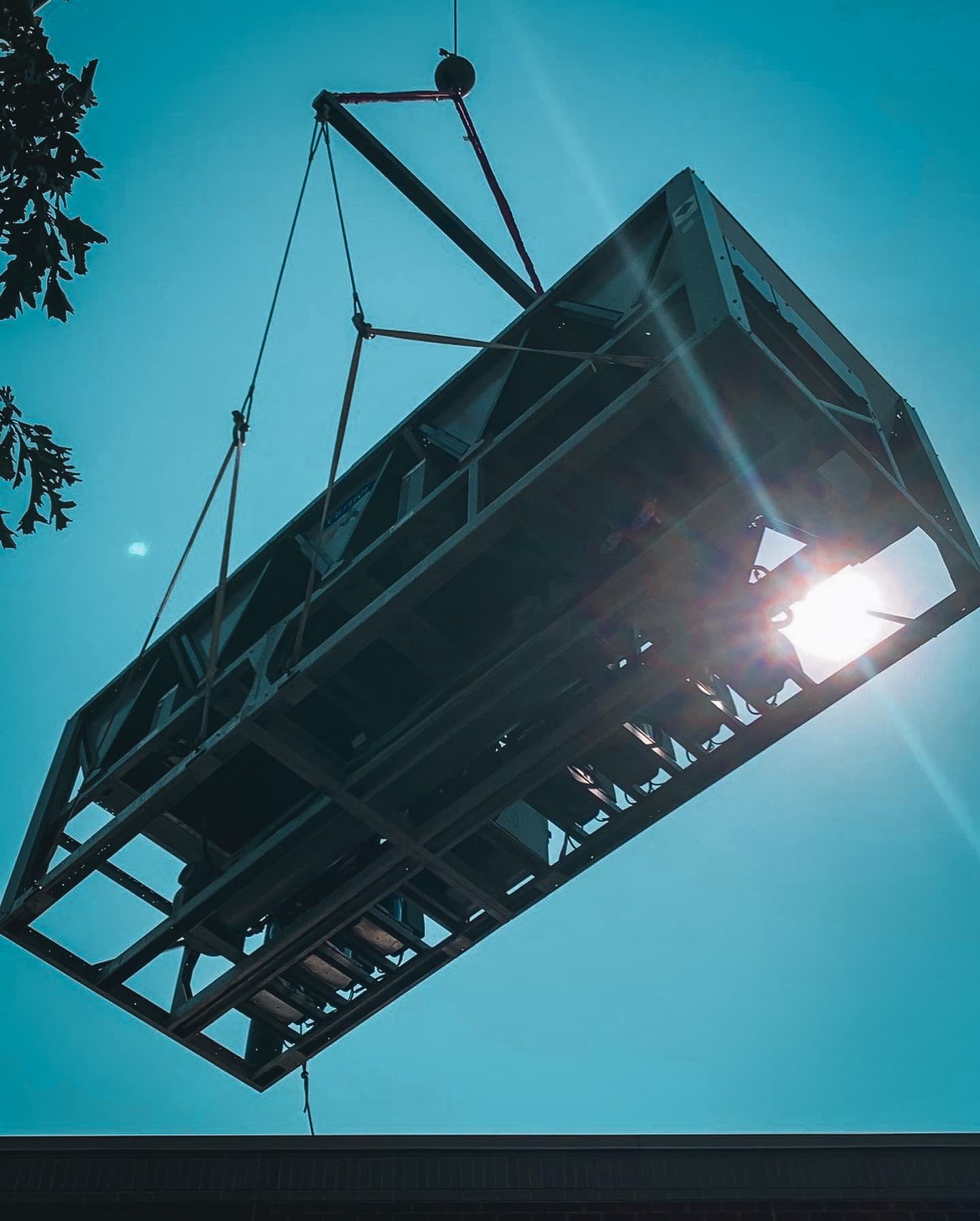 Large industrial HVAC unit suspended by crane cables against the sky during rooftop installation.